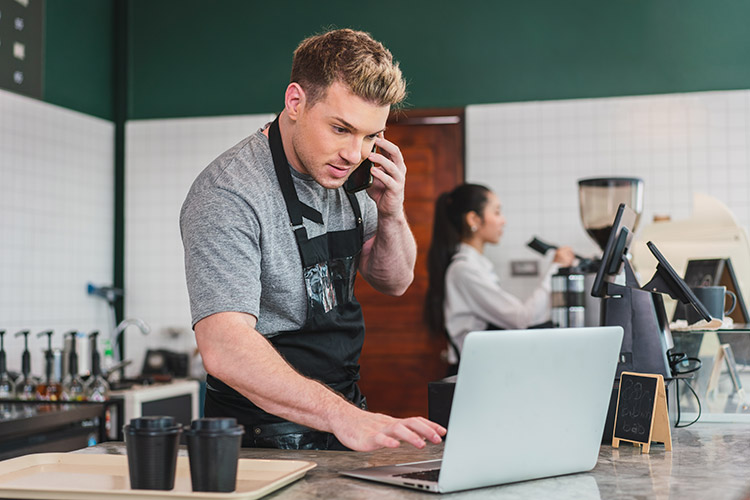 Cafe Owner Barista Receives Order From Customer On Smartphone While Using Computer At Coffee