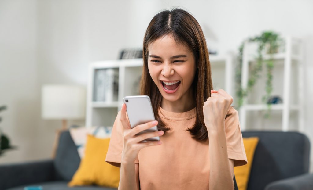 woman holding smartphone with fists clenched celebrating victory expressing